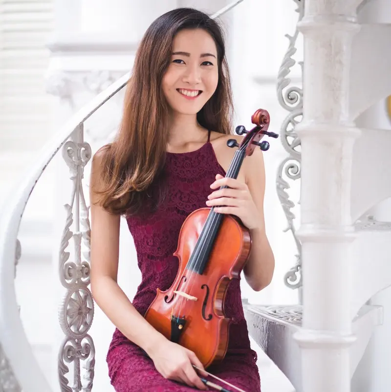 Smiling violinist in a burgundy dress seated with her violin on a white staircase