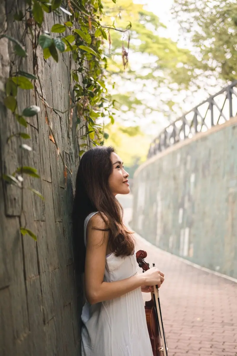 Violinist in a white dress holding her violin against a leafy garden wall in Singapore